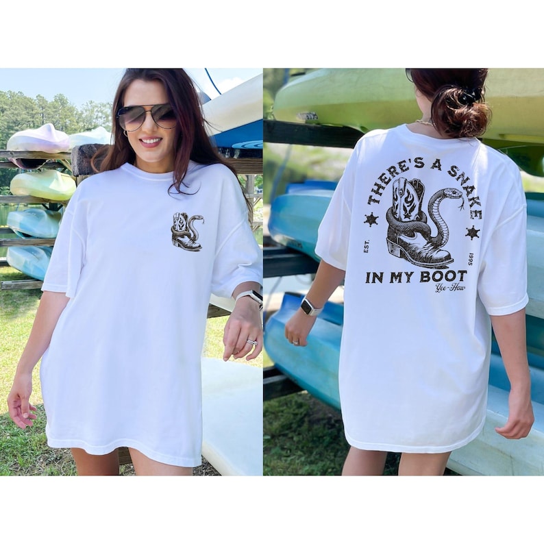 May include: White t-shirt with a graphic of a snake coiled around a cowboy boot. The text on the back of the shirt reads "THERE'S A SNAKE IN MY BOOT" with additional text and stars. The shirt is worn by a person.