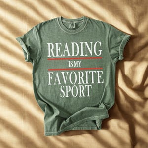 May include: A sage green t-shirt with the text "READING IS MY FAVORITE SPORT" in white, with red lines separating the phrases. The shirt has rolled-up sleeves and a crew neck.