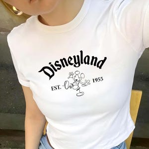 May include: White t-shirt featuring the word "Disneyland" in a stylized font, with "EST. 1955" and a Mickey Mouse graphic. The shirt has a crew neck and short sleeves.