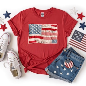 May include: A red short-sleeved t-shirt featuring a central graphic of an American flag composed of various fabric patterns including gingham, floral, and polka dots. The shirt is surrounded by white, red, and blue stars, white sneakers, and denim shorts.