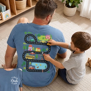 May include: A blue short-sleeved t-shirt with a road map design on the back, featuring houses, trees, a school bus, and a pond. The shirt has the text "Tired Dads Club" on the front. A child is playing with toy cars on the shirt.