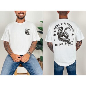May include: White t-shirt with a graphic of a cowboy boot and snake. The front of the shirt has a small graphic of the boot and snake. The back of the shirt has the text "THERE'S A SNAKE IN MY BOOT" with a boot and snake graphic.