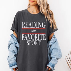 May include: Dark gray t-shirt with the text "READING IS MY FAVORITE SPORT" in white, with red lines above and below the text. The shirt has rolled-up sleeves and is worn with a denim jacket.