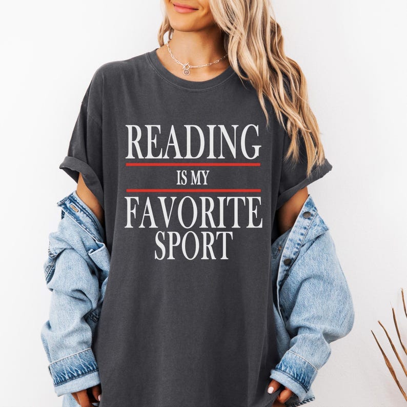 May include: Dark gray t-shirt with the text "READING IS MY FAVORITE SPORT" in white, with red lines above and below the text. The shirt has rolled-up sleeves and is worn with a denim jacket.