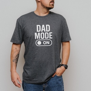 May include: Dark gray t-shirt with the words "DAD MODE ON" printed in white. The design includes an on/off switch graphic. The shirt is short-sleeved and made of a soft material.