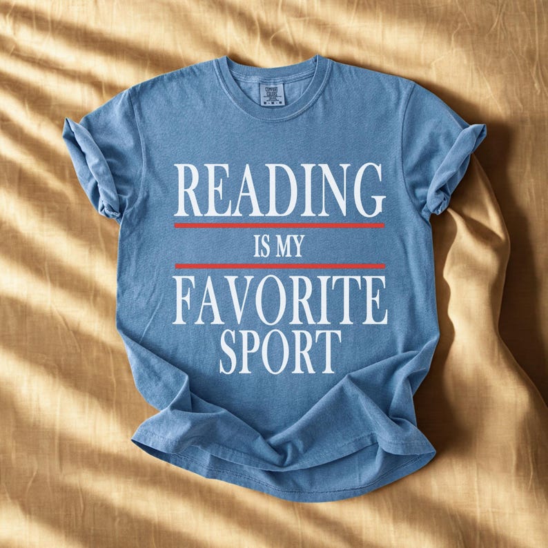 May include: A blue t-shirt with the text "READING IS MY FAVORITE SPORT" in white. The text is separated by two red lines. The shirt is folded and lies on a tan surface.