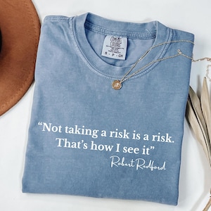 May include: A blue Comfort Colors t-shirt with the quote "Not taking a risk is a risk. That's how I see it" by Robert Redford. A gold necklace with a pendant rests on the shirt. The shirt is folded and has a tag.