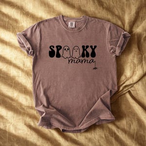 May include: A mauve-colored t-shirt with the words "Spooky Mama" in black script and a spider graphic. The word "Spooky" is in a bold, retro font, with two ghost illustrations. The shirt is folded on a golden-yellow surface.