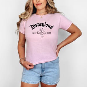 May include: A light pink t-shirt with the word "Disneyland" in black script. Below the word is a graphic of Mickey Mouse and the text "EST. 1955". The shirt is short-sleeved and is being worn with blue jean shorts.