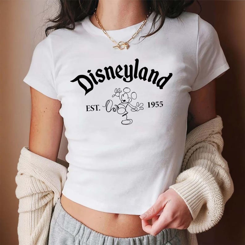 May include: White crop top featuring the word "Disneyland" in a black script font, with "EST. 1955" and a Mickey Mouse graphic. The shirt is a short-sleeved, crew neck style.