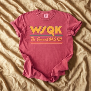 May include: A red t-shirt with rolled-up sleeves featuring the text "WSQK The Squawk 94.5 FM" in yellow, green, and orange. The design includes a lightning bolt graphic. The shirt is displayed on a tan surface.