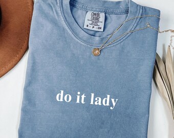 Puede incluir: Una camiseta azul Comfort Colors con la frase "do it lady" en letras blancas. Un collar dorado con un colgante descansa sobre la camiseta doblada. Un sombrero marrón es parcialmente visible. La camiseta tiene una etiqueta pequeña.