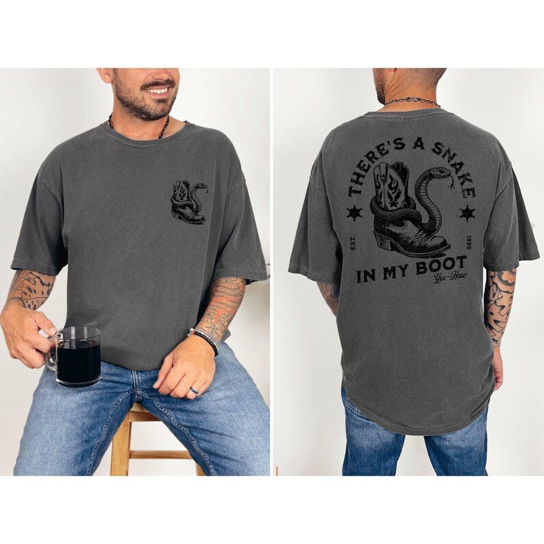 May include: Dark gray t-shirt with a graphic of a cowboy boot and snake. The back of the shirt features the text "THERE'S A SNAKE IN MY BOOT" in a circular design. The shirt has a relaxed fit and short sleeves.