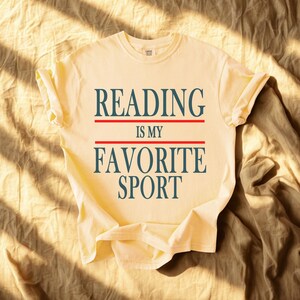 May include: A light yellow t-shirt with the text "READING IS MY FAVORITE SPORT" in navy blue letters, separated by red lines. The shirt is folded, and the sleeves are cuffed. The shirt is on a tan fabric background.