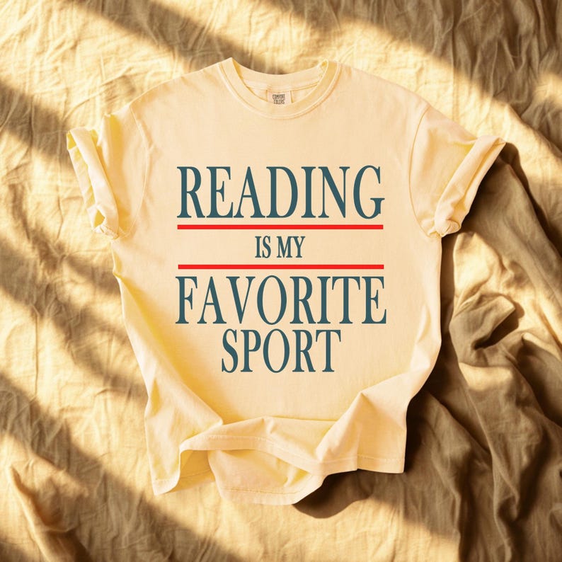 May include: A light yellow t-shirt with the text "READING IS MY FAVORITE SPORT" in navy blue letters, separated by red lines. The shirt is folded, and the sleeves are cuffed. The shirt is on a tan fabric background.