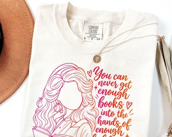 Może przedstawiać: Kremowy t-shirt z grafiką przedstawiającą osobę z długimi, falowanymi włosami czytającą książkę. Tekst brzmi: "You can never get enough books into the hands of enough children". Wzór wykorzystuje gradient kolorów różowego, pomarańczowego i czerwonego.