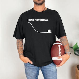 Puede incluir: Camiseta negra con el texto blanco "I HAD POTENTIAL" sobre un gráfico de una bola rodando por una línea curva. La persona de la imagen sostiene un balón de fútbol americano marrón y blanco.