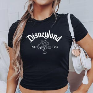 May include: Black t-shirt with the word "Disneyland" in white script, along with a Mickey Mouse outline and the text "EST. 1955". The shirt is a crew neck style.
