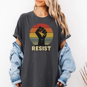 May include: Dark gray t-shirt featuring a retro design of a raised fist inside a striped circle with red, orange, yellow, and green hues. The word "RESIST" is printed in a bold, sans-serif font below the graphic. The shirt has rolled-up sleeves.