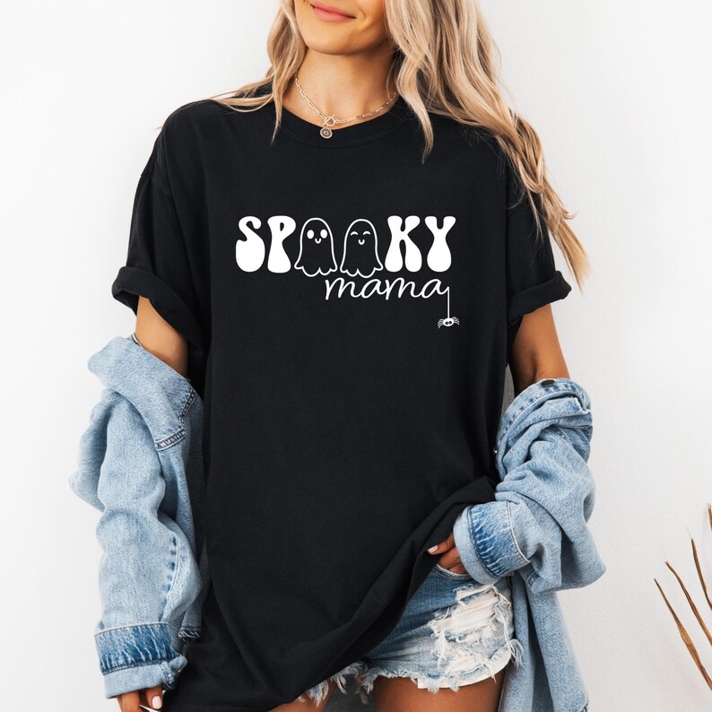 May include: Black t-shirt with the words "Spooky Mama" in white, featuring two ghost illustrations. A small spider graphic hangs from the word "mama". The shirt is a casual, oversized fit.
