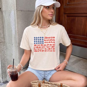May include: A cream-colored short-sleeve t-shirt featuring a graphic design of the American flag composed of blue stars and red illustrations of various sea creatures, including crabs, lobsters, and fish, arranged in a rectangular grid pattern.