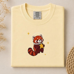 May include: A pale yellow t-shirt featuring an embroidered design of a red panda wearing a space helmet and holding a yellow star. The design includes small yellow star accents. The inner neck label reads Comfort Colors, S-P-CH.
