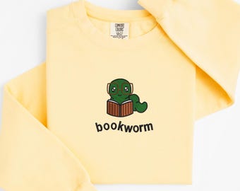 May include: A light yellow sweatshirt with a cartoon graphic of a green worm wearing glasses and reading a book. The word "bookworm" is printed below the graphic in black. The sweatshirt has long sleeves and a crew neck.