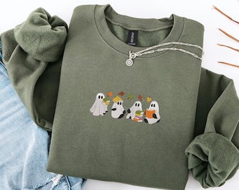 May include: An olive green sweatshirt featuring an embroidered design of four cartoon ghosts reading books. The ghosts are in different poses, with autumn leaves above. A silver necklace with a pendant is draped across the sweatshirt.