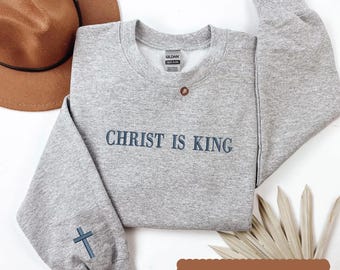 May include: A heather gray sweatshirt with the text "CHRIST IS KING" in navy blue. A navy blue cross is embroidered on the left sleeve. The sweatshirt has a crew neck and a brown button detail. A brown hat is in the background.
