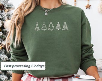 May include: A dark green sweatshirt with a crew neck featuring a white line drawing of five Christmas trees. The text "Fast processing 1-2 days" is in a white box in the lower left corner. The person is wearing red and white plaid pajama pants.