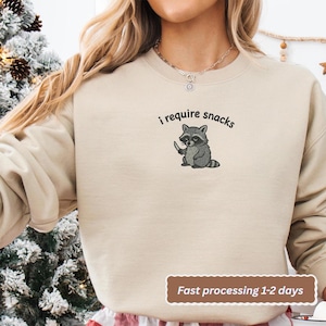 May include: Beige sweatshirt with the text "i require snacks" above a cartoon raccoon holding a knife. The raccoon is gray and black. The sweatshirt is a neutral color and has a crew neck.