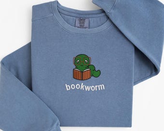 May include: A blue long-sleeve sweatshirt featuring an embroidered design of a green worm wearing glasses and reading a book. The word "bookworm" is embroidered below the graphic in white.