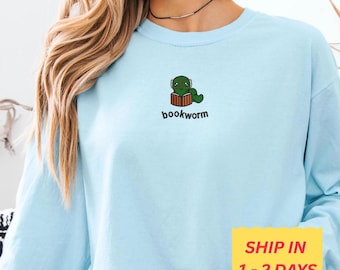 May include: A light blue sweatshirt featuring a green cartoon bookworm reading a book. The word "bookworm" is printed below the graphic. The long-sleeved shirt is made of a soft material.