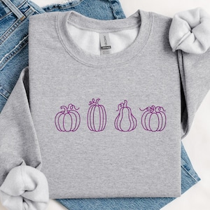 May include: A grey sweatshirt with a row of four purple pumpkin outlines. The pumpkins have different shapes and sizes, with curly stems. The sweatshirt is folded on top of a pair of blue jeans.