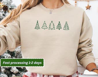 May include: A beige sweatshirt featuring a green line drawing of five Christmas trees. The text "Fast processing 1-2 days" is in a green box at the bottom. The sweatshirt is worn by a person.
