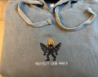May include: A faded blue-gray hooded sweatshirt with a drawstring hood. The center features an embroidered graphic of a black winged creature with red eyes carrying a tan, furry figure. Below the graphic, the text reads PROTECT OUR PARKS.