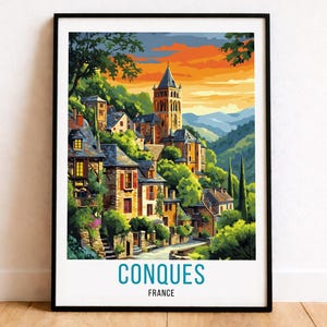 May include: A framed art print of Conques, France, featuring a colorful illustration of a village nestled on a hillside. The artwork showcases buildings with red-tiled roofs, a tall church tower, and lush green trees under a vibrant sunset sky.