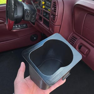 Op de afbeelding: Een zwarte, rechthoekige, 3D-geprinte bekerhouder-inzet met een ronde binnenholte en een platte, brede bovenrand. Het object heeft een getextureerde oppervlakteafwerking en een klein uitstekend lipje aan één kant voor in de console.