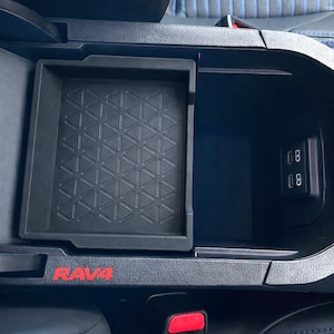 Op de afbeelding: Een zwarte kunststof middenconsole-organizer met een geometrisch driehoekspatroon op de bodem, geïnstalleerd in een opbergvak van een voertuig. De console bevat de rode tekst RAV4 en twee USB-C-oplaadpoorten op de binnenwand.