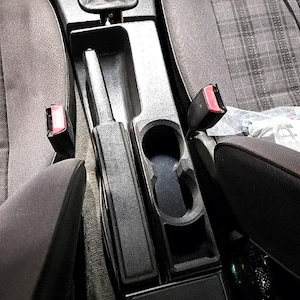 Peut inclure: Un insert de console centrale en plastique noir doté d'une découpe pour le levier de frein à main et de deux porte-gobelets circulaires intégrés. La console est placée entre deux sièges de voiture en tissu gris avec des boucles de ceinture rouges.