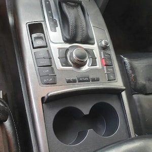 Op de afbeelding: Een donkergrijze middenconsole met een zwarte lederen versnellingspook, een centrale bedieningsknop en diverse knoppen met de labels NAV, INFO, CAR, SETUP, RETURN en TEL. Onder de bedieningselementen bevinden zich twee zwarte bekerhouders.