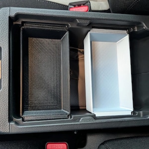Op de afbeelding: Een zwarte kunststof middenconsole-organizer met twee rechthoekige opbergvakken. Het ene vak is zwart en het andere is lichtgrijs, beide ontworpen om in de diepe opbergruimte van de middenconsole van een voertuig te passen.