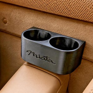 May include: A black dual cup holder featuring the word Miata embossed on the front surface. The accessory is designed with two circular openings and a smooth, matte finish, positioned securely within a tan leather vehicle interior setting.