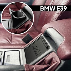 Może przedstawiać: Czarny plastikowy uchwyt na kubek do konsoli środkowej pojazdu BMW E39. Wkładka posiada prostokątną podstawę z widocznym na zdjęciu napisem BMW E39 oraz okrągły otwór na napoje, pokazany w pozycji zamkniętej i otwartej.