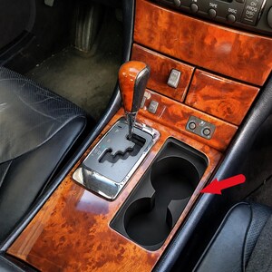 Peut inclure: Une console centrale de voiture avec une garniture en grain de bois, un levier de vitesse avec un pommeau assorti en grain de bois et un porte-gobelet double en plastique noir. Une flèche rouge pointe vers la zone du porte-gobelet.