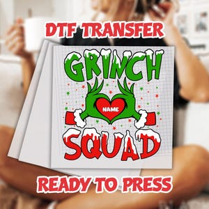 May include: A stack of white DTF transfer sheets featuring a Grinch-themed design. The design includes the words "Grinch Squad" in red and green, with a heart-shaped hand gesture. The text "Ready to Press" is also included.