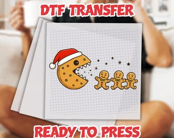 May include: DTF TRANSFER and READY TO PRESS text over a graphic of a cookie wearing a Santa hat, eating gingerbread men. The design is on a grid paper background. The cookie has orange dots and a white mouth.