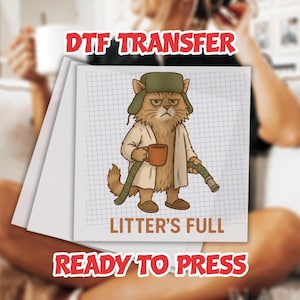 May include: DTF transfer featuring a grumpy cat cartoon wearing a hat and coat, holding a mug and a hose. The image includes the text "LITTER'S FULL" and "READY TO PRESS". The background is a grid pattern.