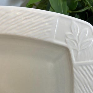 May include: Cream-colored rectangular serving dish with a textured border. The border features a leaf design in one corner and a diagonal pattern on the sides. The dish is made of ceramic and has a smooth, glossy surface.