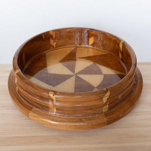 May include: A round wooden bowl with a geometric pattern of light and dark wood. The bowl is made of multiple pieces of wood that are inlaid together to create the design.